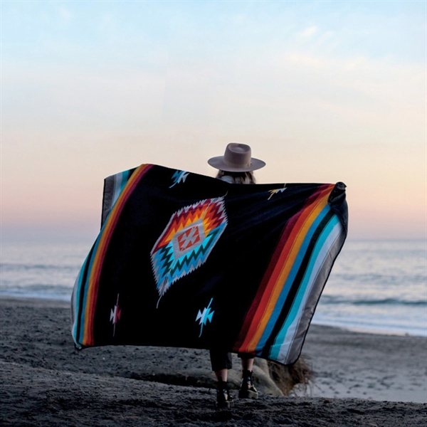 woman holding out branded sustainable merch slowtide blanket with unique colored pattern on the beach